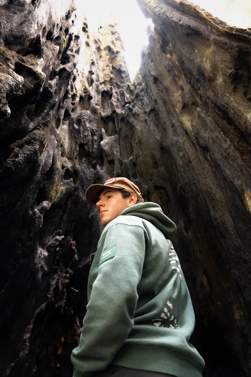 A man wearing a green hoodie and a tan cap standing among tall dark rocks inside a cave or crevice, with light shining from above.