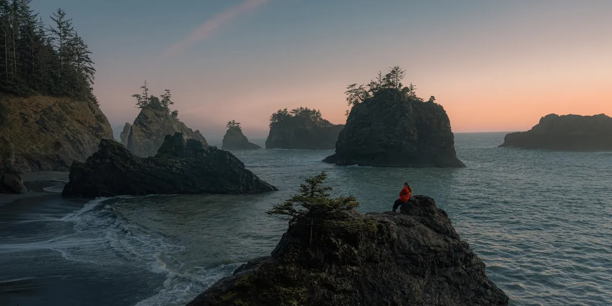 A person sitting on a rock near the ocean with large rocky islands covered in trees in the background during sunset.